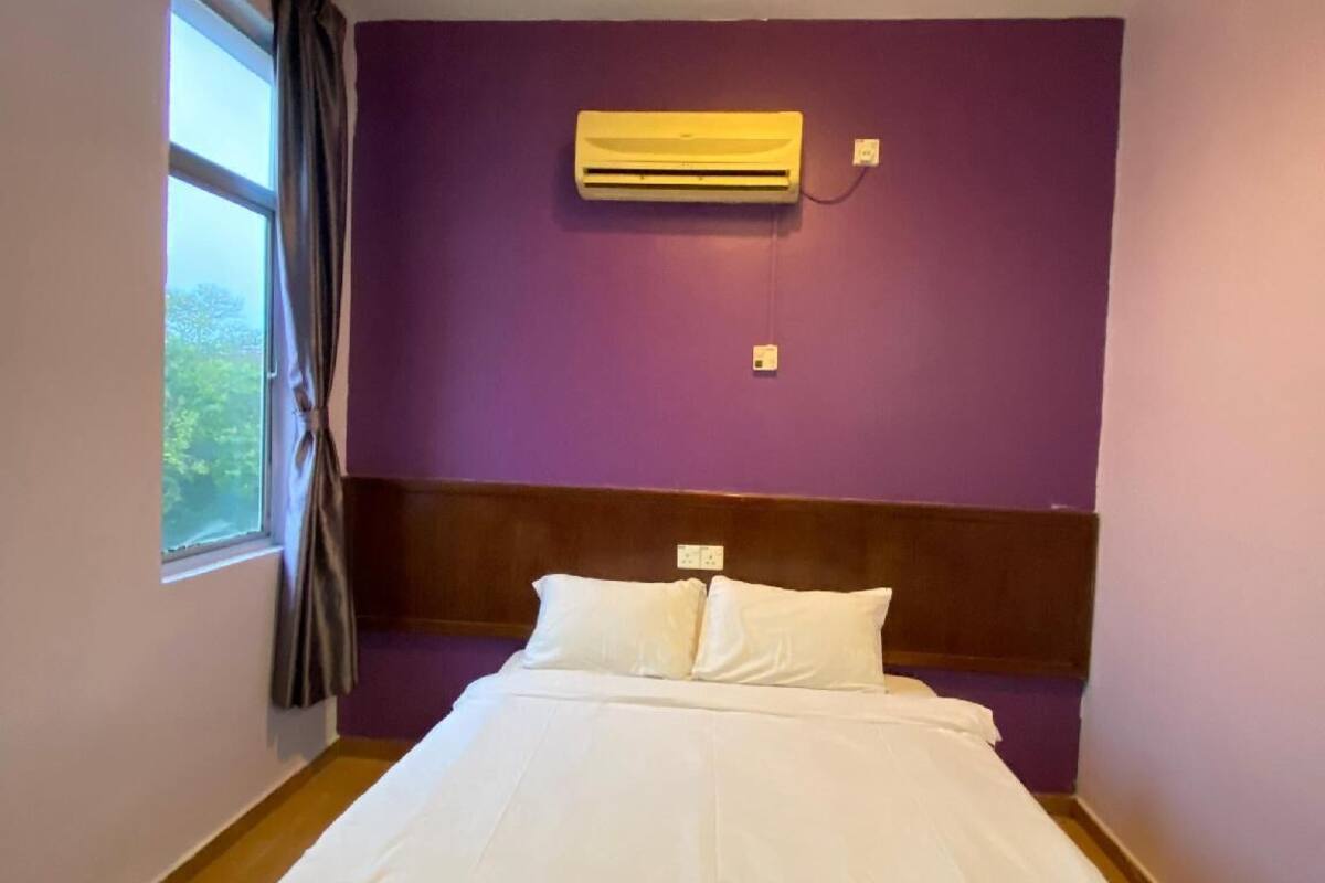 Habitación de hotel con una cama, un equipo de aire acondicionado en la pared, una ventana con cortinas y un cabecero de madera.