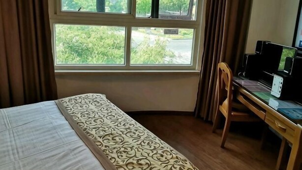 A hotel room with a bed, a desk with a computer, and a window with a view of buildings and greenery.