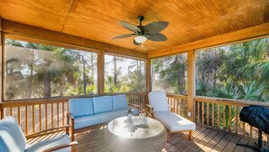 A covered outdoor deck with wooden flooring, a ceiling fan, a round table with a bottle, a blue sofa, and a white lounge chair. - Marsh View Treehouse Retreat on Fripp Island (Fripp Island)