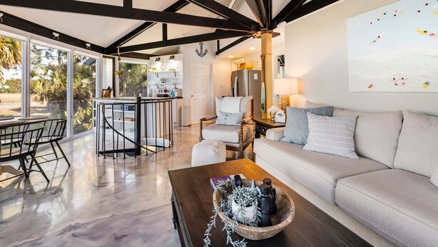 A spacious living area with a large window, a wooden coffee table, a sofa, and a kitchen in the background. - Marsh View Treehouse Retreat on Fripp Island (Fripp Island)