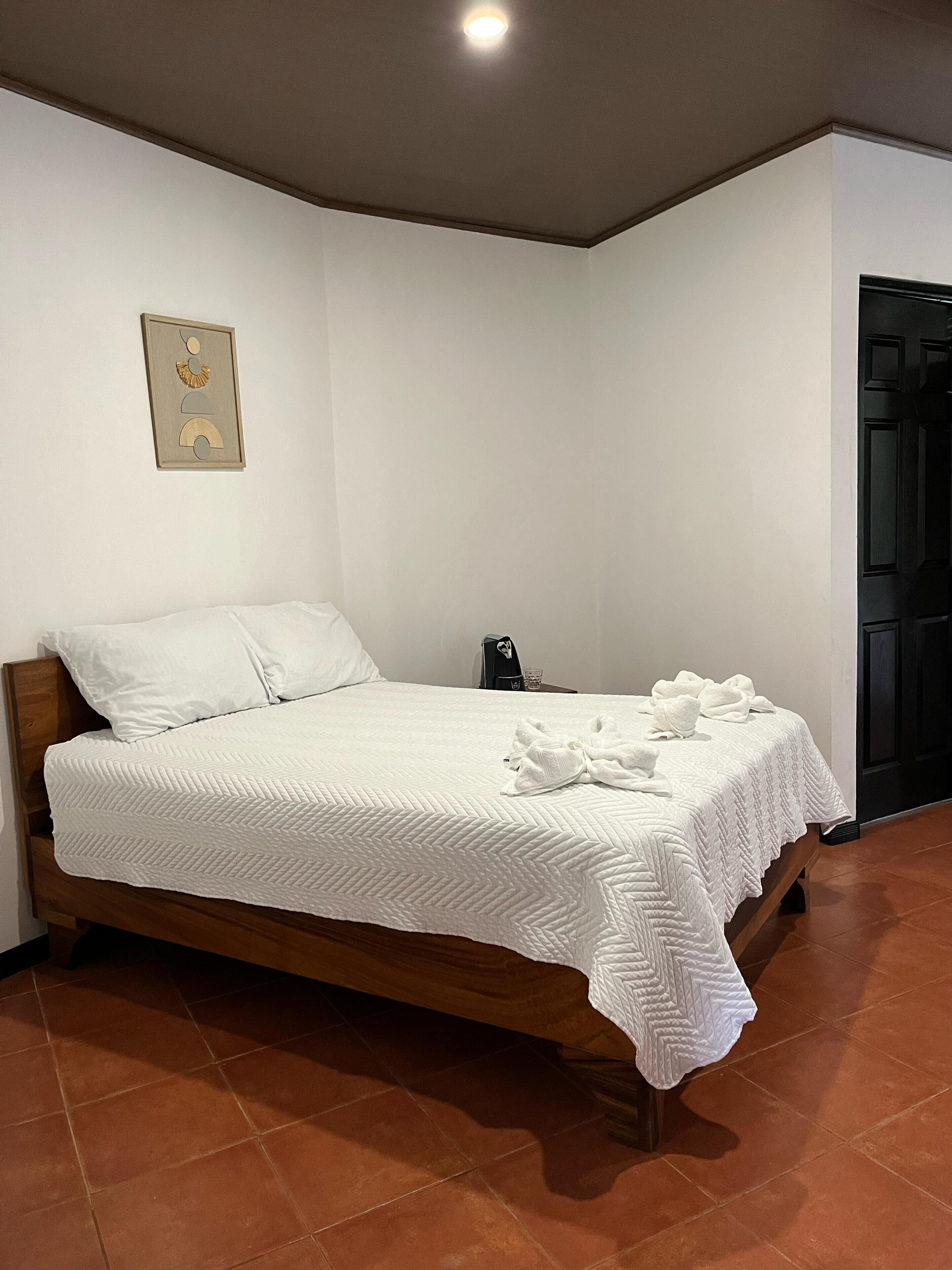 A neatly made bed with white linens and a quilted blanket, a bedside table with a phone and a glass, and a framed picture on the wall.