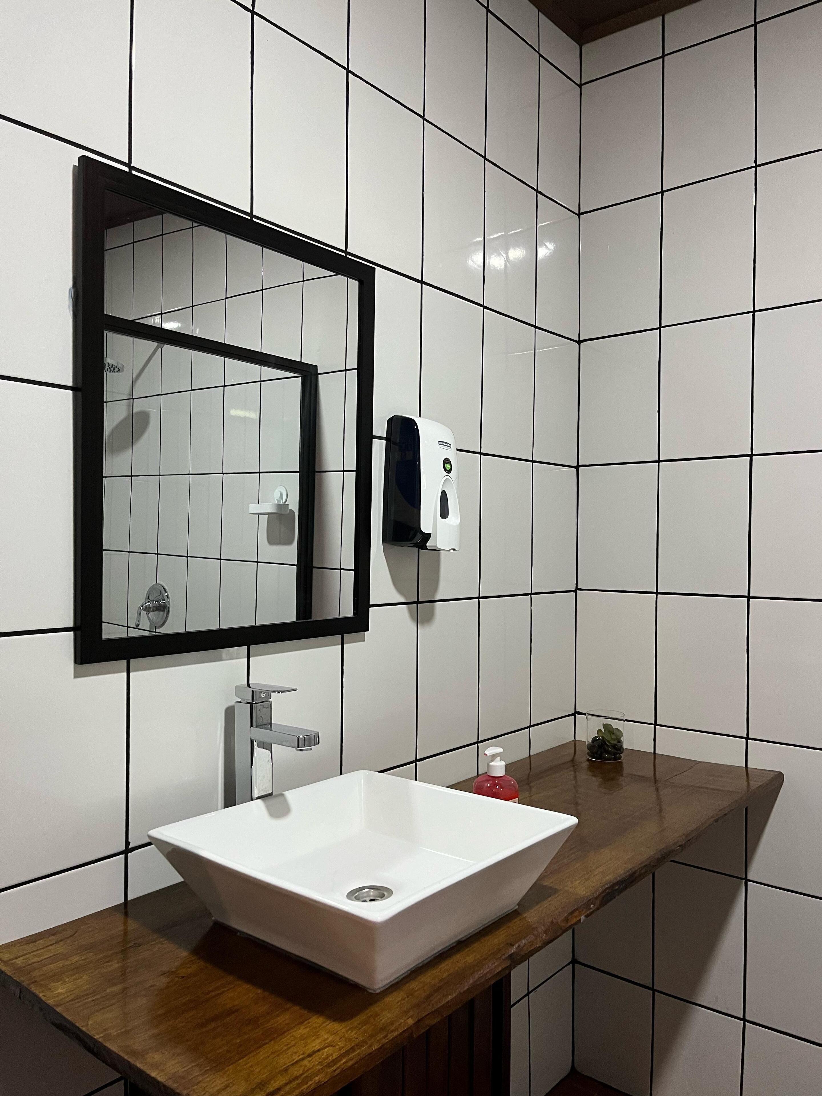 A bathroom with white tiled walls, a rectangular mirror with a black frame, a white sink with a wooden countertop, a soap dispenser, and a small glass container with a plant.