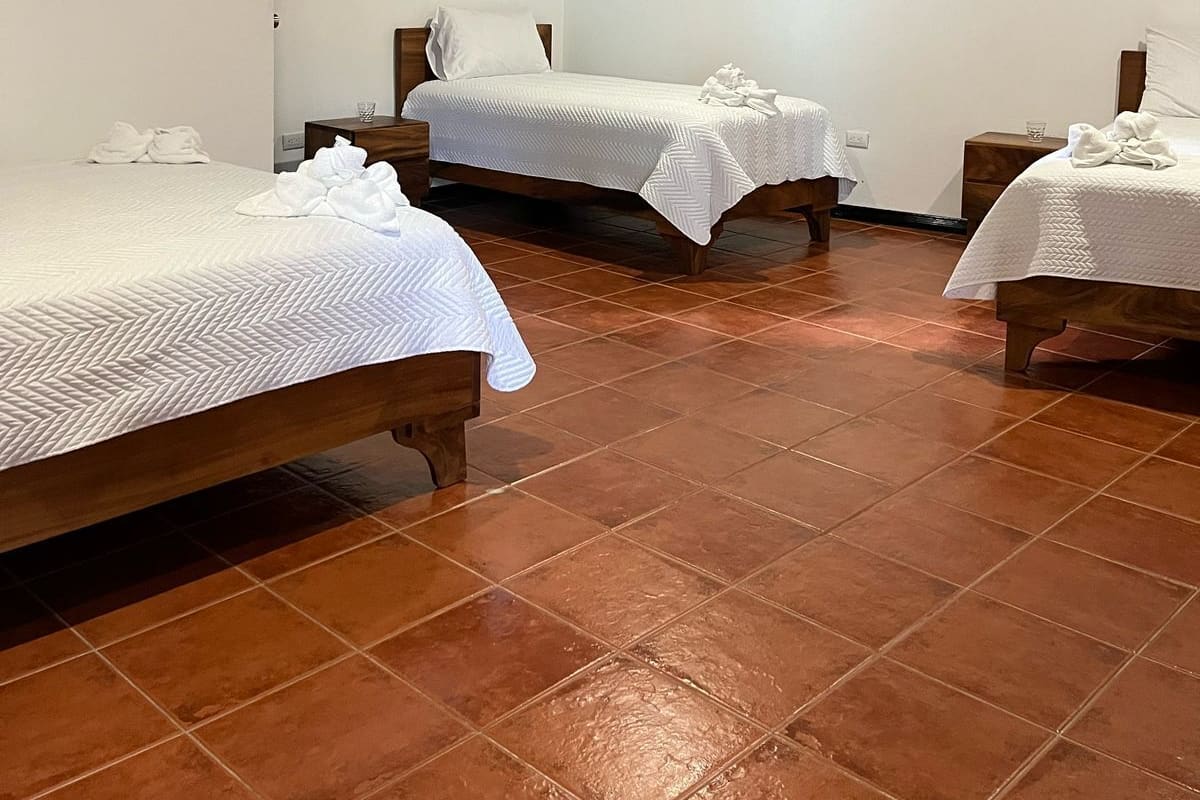 A hotel room with three beds, each with white bedding and a wooden headboard. The room has a tiled floor and a white wall with an air conditioning unit mounted above the beds.