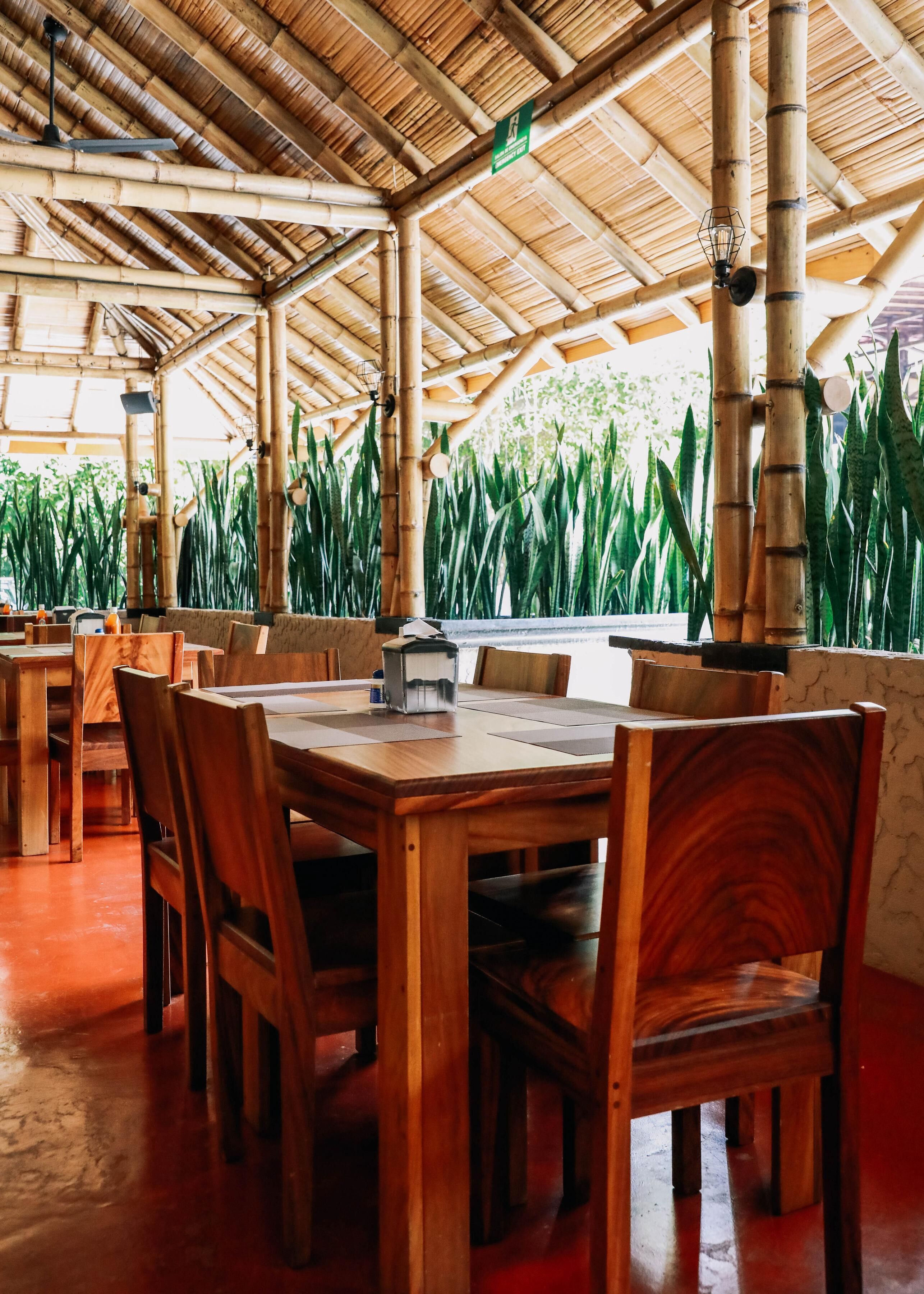 A bamboo-paneled room with wooden tables and chairs, a ceiling fan, and a view of green plants.