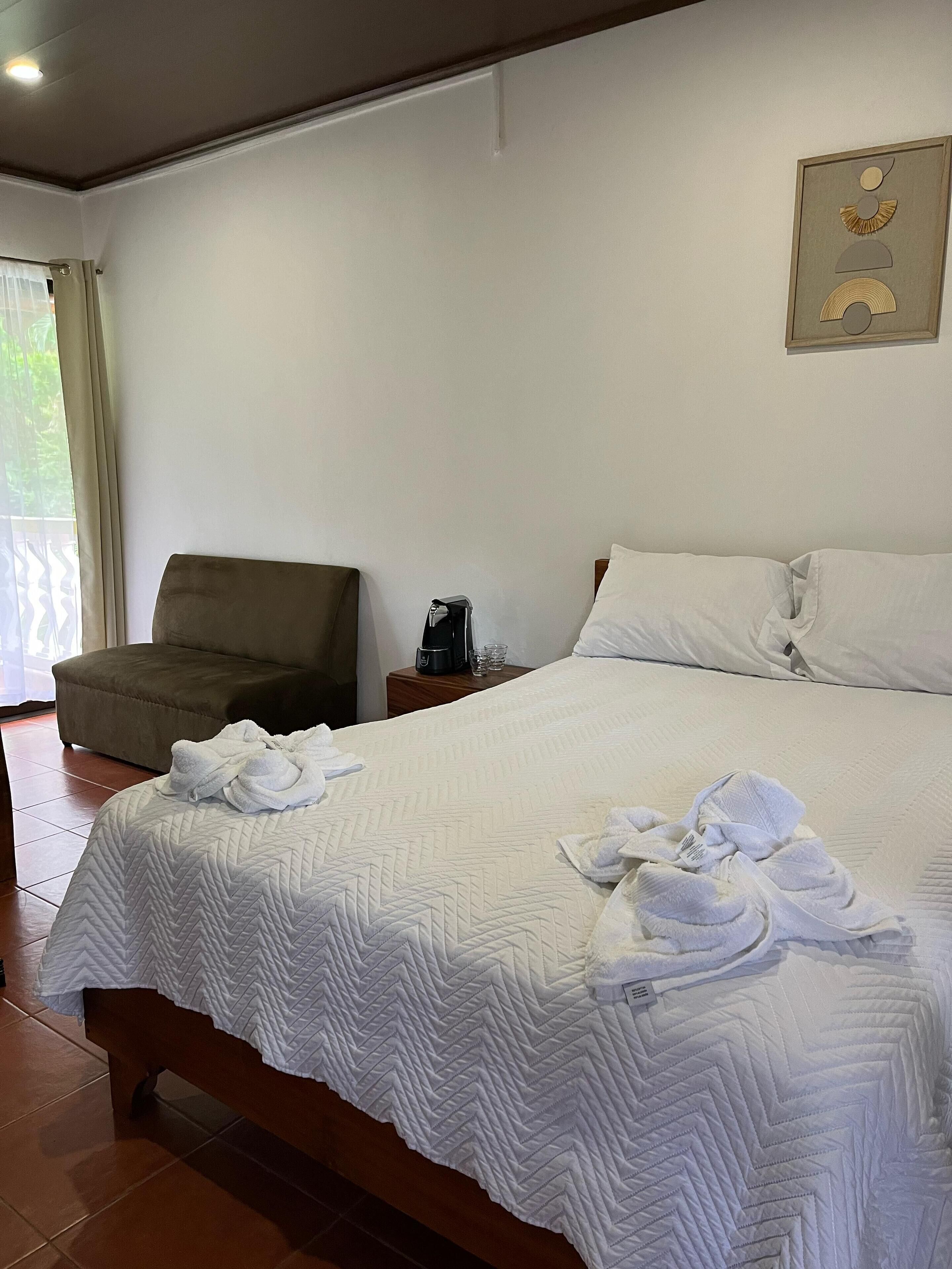 A neatly made bed with white linens and towels, a brown armchair, a bedside table with a coffee maker, and a framed artwork on the wall.