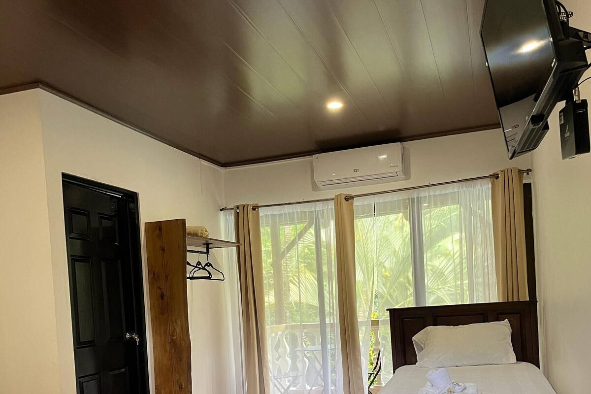 A hotel room with a bed, a chair, a wooden pillar, a black door, and a window with curtains.