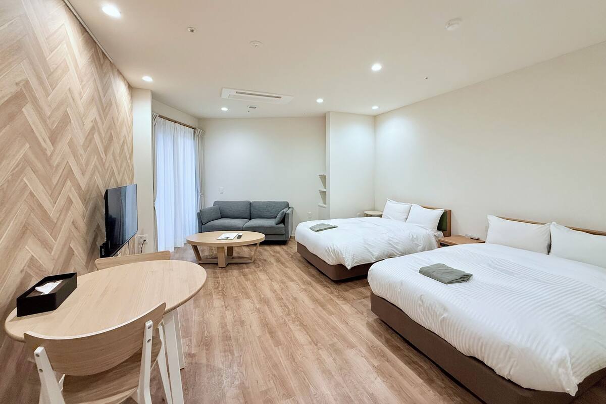 Una habitación de hotel moderna con suelo de madera en patrón de espiga, un sofá gris, dos camas individuales, una mesita redonda y un televisor.