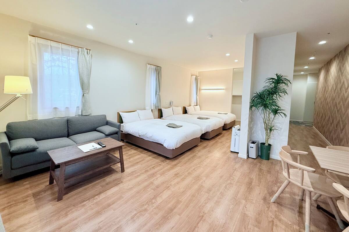Una habitación de hotel moderna con sofá, cama con dos almohadas, mesita de noche, lámpara, planta y suelo de madera.