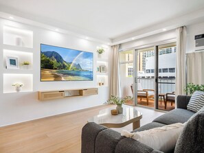 A modern living room with a large flat-screen TV, wooden flooring, a grey sofa, and a glass door leading to a balcony with a city view. - 2 Bedroom 2 Bath + Den 5 beds Partial Ocean View + 1 Parking 2min from the Beach (Honolulu)