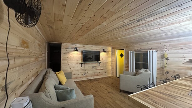L’intérieur chaleureux d’un chalet avec un plafond en bois, un canapé, un téléviseur fixé au mur et une table à manger avec des chaises.