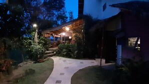 Ein Weg führt zu einem überdachten Außenbereich mit Holz-Pergola, umgeben von Grünpflanzen und einem weißen Gebäude.