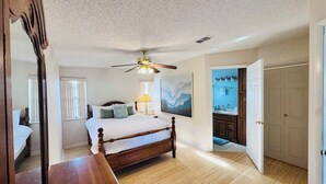Camera da letto con un letto in legno, una ventola a soffitto, un quadro e un bagno visibile attraverso una porta aperta.