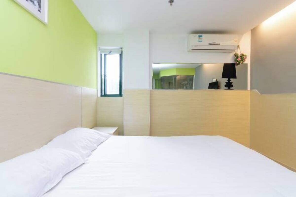A hotel room with a bed, a window, a wall-mounted air conditioner, and a small table with a lamp.