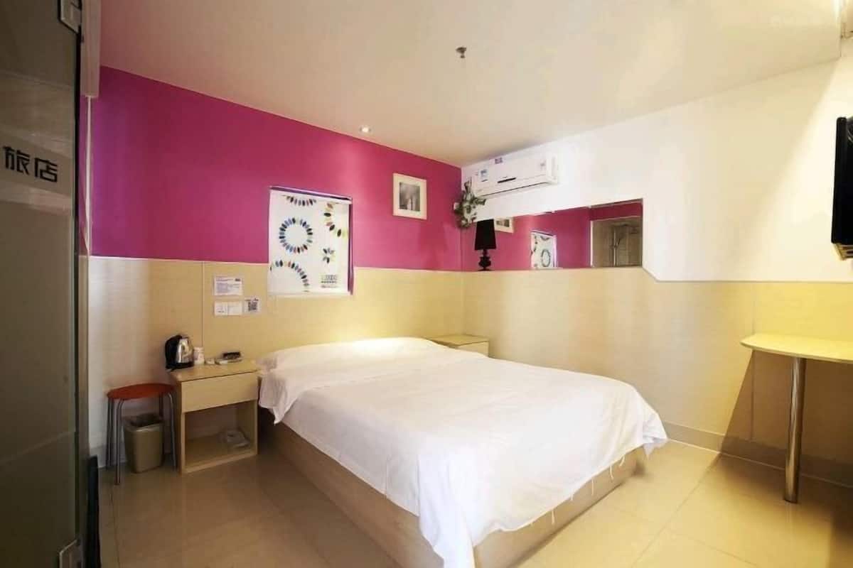 A hotel room with a bed, a desk, a chair, a trash bin, and an air conditioning unit.