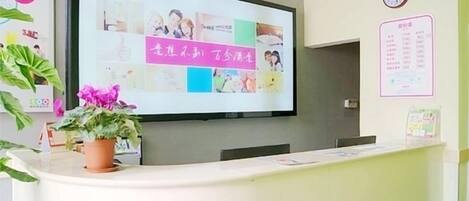 A reception desk with a flat-screen TV mounted on the wall displaying a presentation, a potted plant, and a wall clock.
