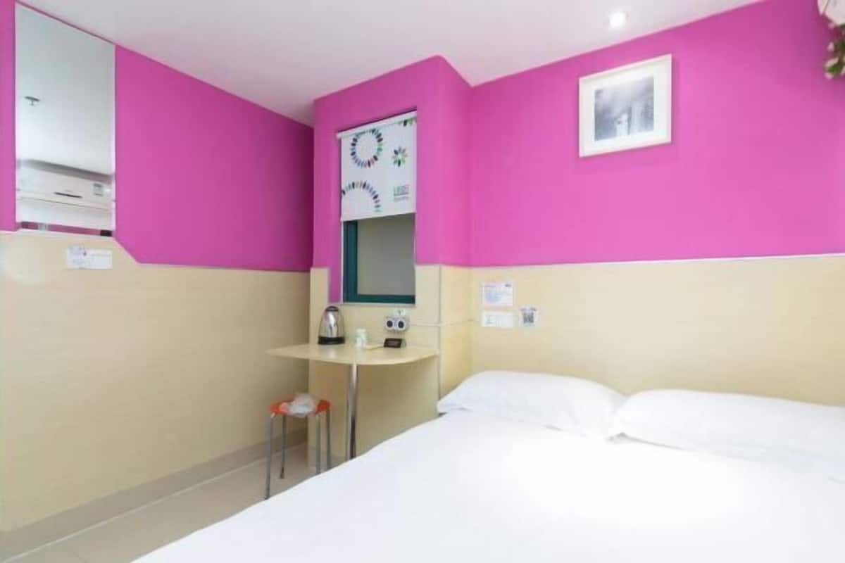 A hotel room with a bed, a small table with a kettle, a chair, a mirror, and a picture on the wall.