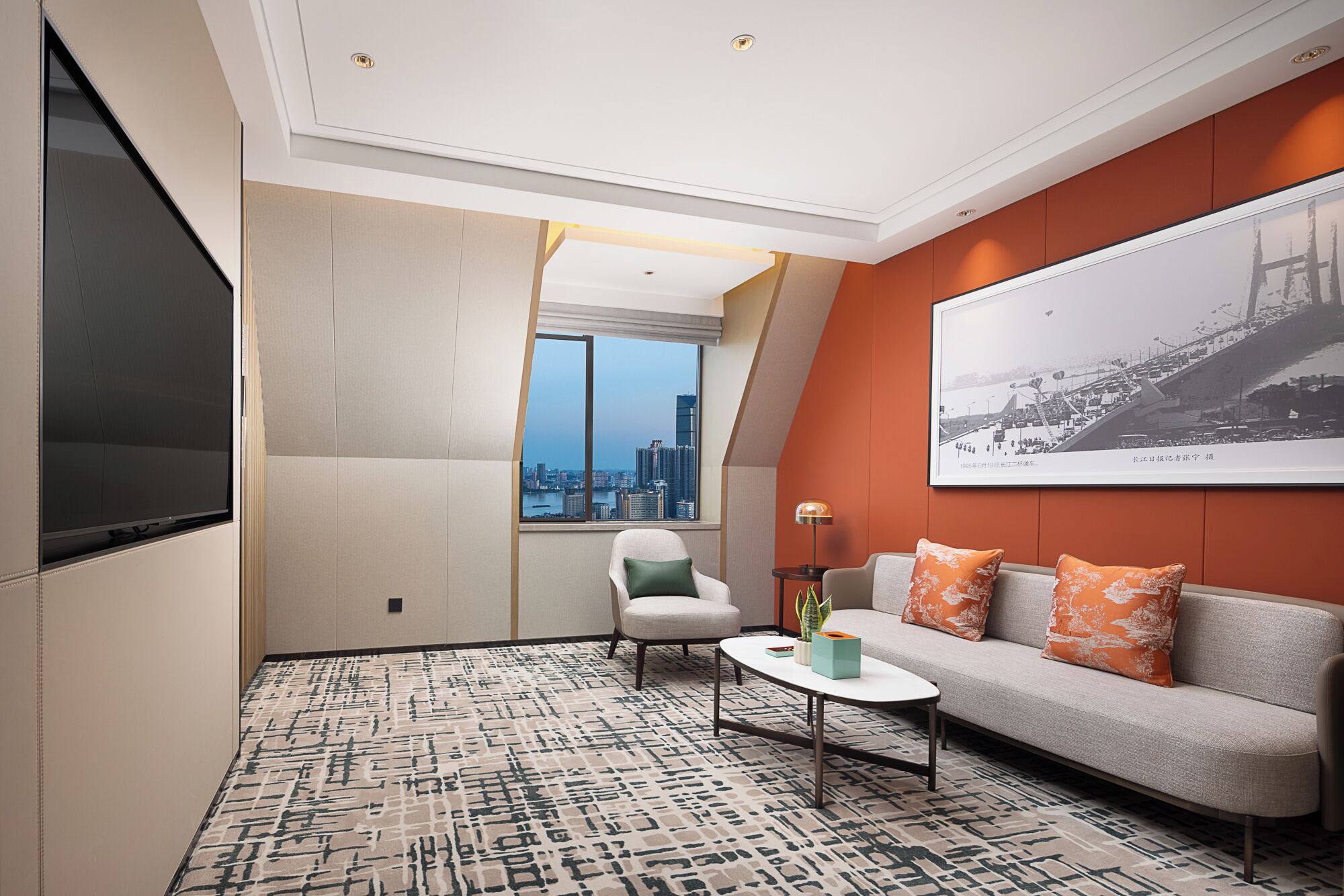 A modern hotel room with a city view, a sofa with orange cushions, a small table, and a large framed picture on the wall.