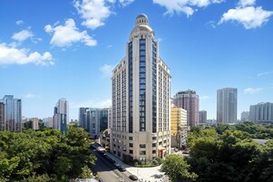 Un edificio alto con una destacada torre de reloj, rodeado de otros edificios altos y vegetación.