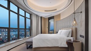Une chambre d’hôtel moderne avec un grand lit, une table de chevet, une lampe et une vue sur le paysage urbain grâce à de grandes fenêtres.