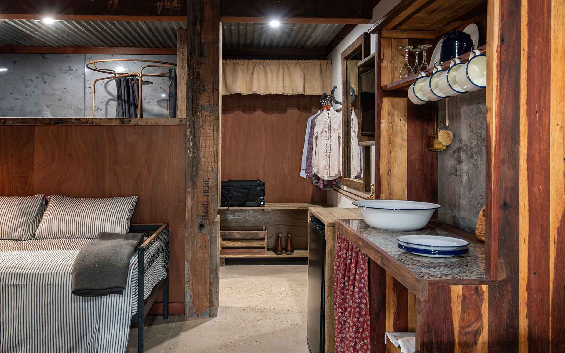 Interior rústico de una cabaña con una cama, un banco de madera, un lavabo y un espejo con objetos colgando.