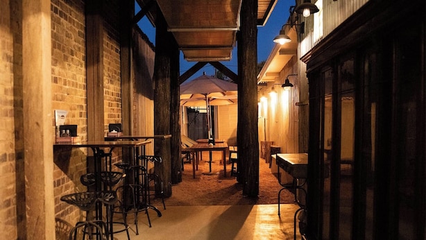 An outdoor corridor with tables and chairs, a brick wall, and a building in the background. - The Staging Post (Longreach)