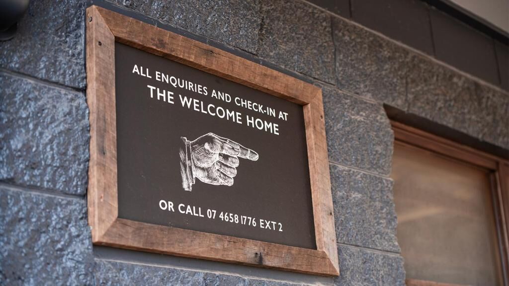 A sign with a pointing hand graphic and text inviting enquiries and check-ins at the welcome home, with a contact number.