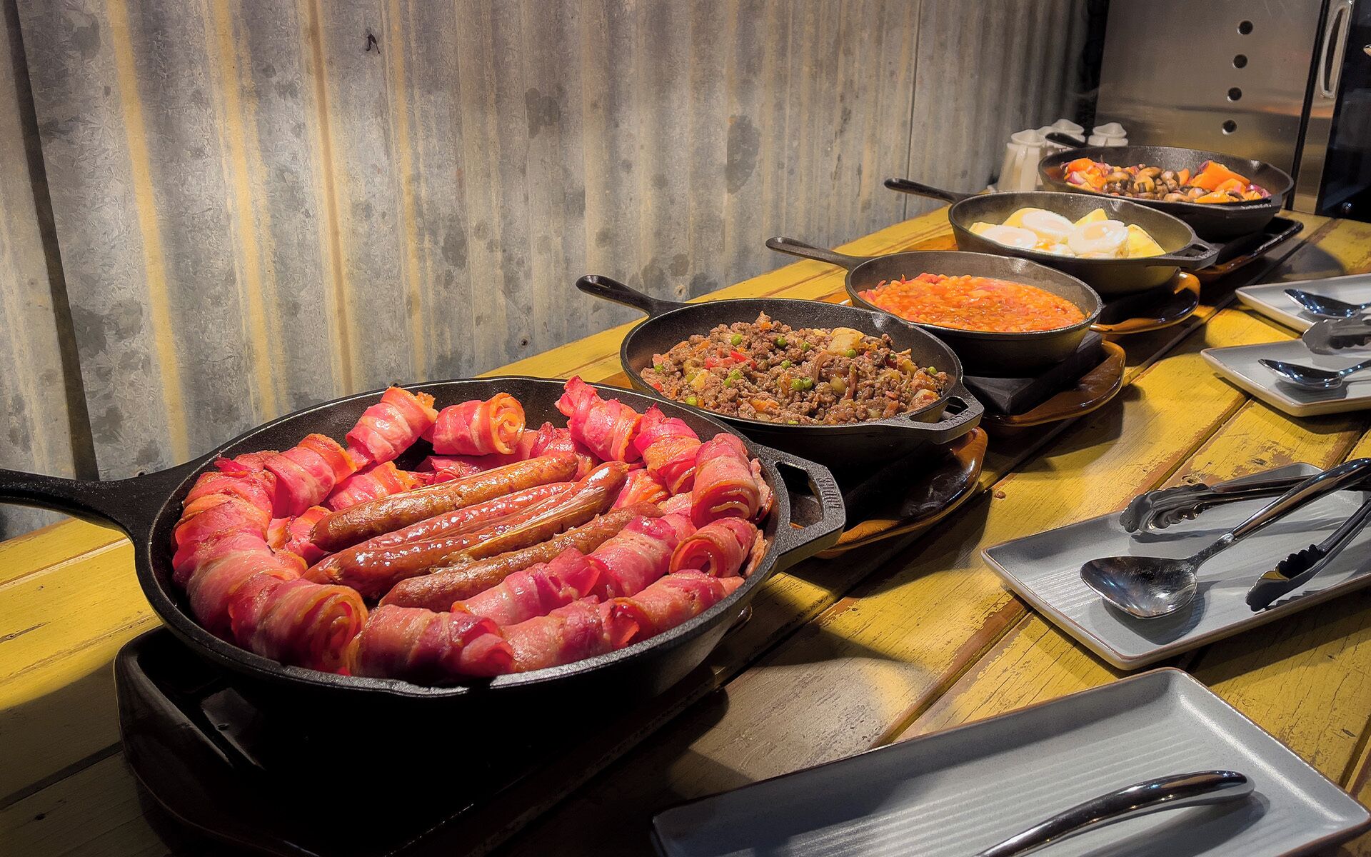Uma mesa com diversos pratos, incluindo salsichas envoltas em bacon, uma frigideira com um prato de carne e uma panela com um prato de legumes.