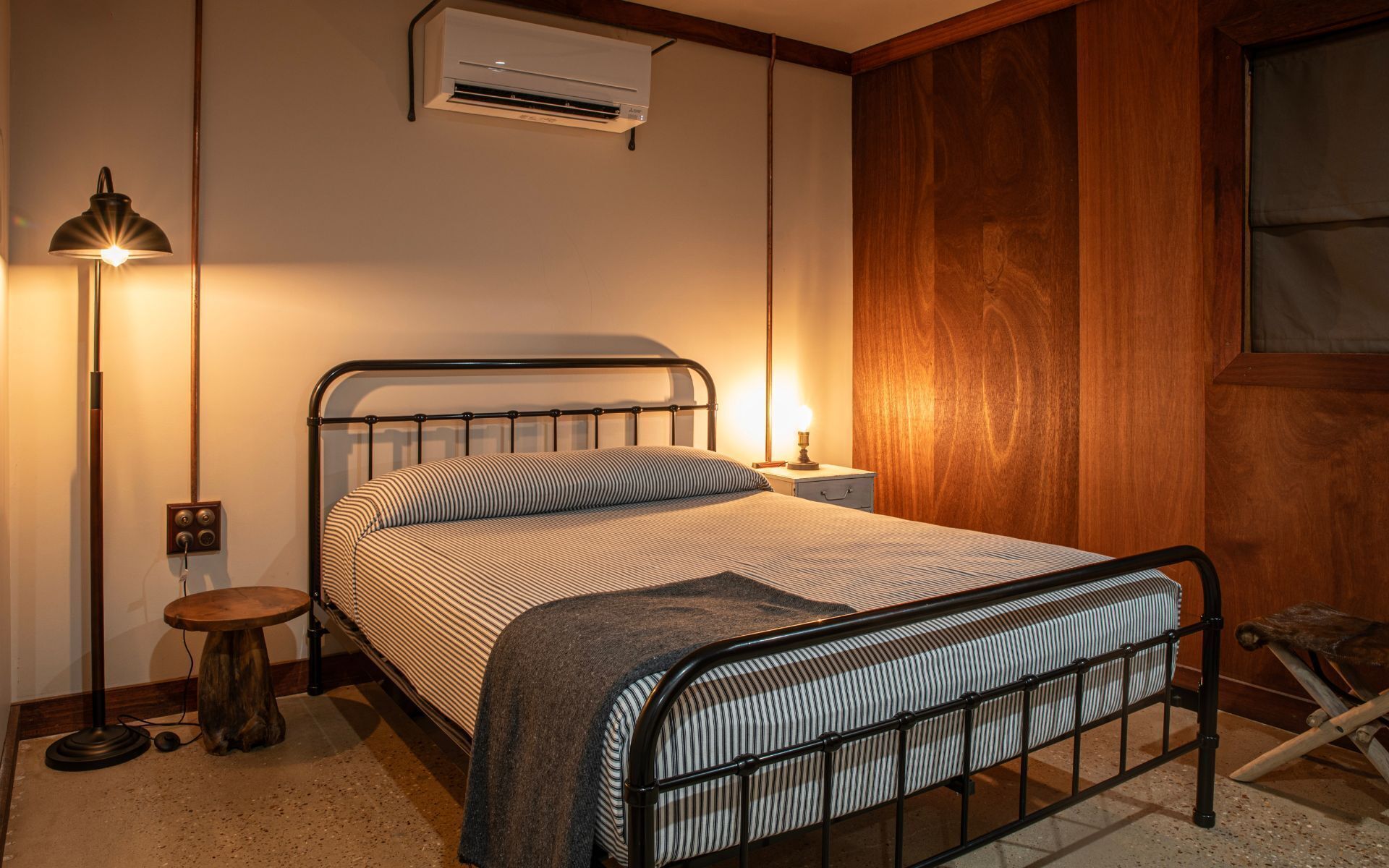 Una cama con estructura metálica, una lámpara de noche, aire acondicionado y un taburete pequeño de madera.