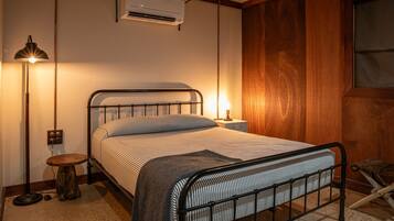 Una cama con estructura metálica, una lámpara de noche, aire acondicionado y un taburete pequeño de madera.