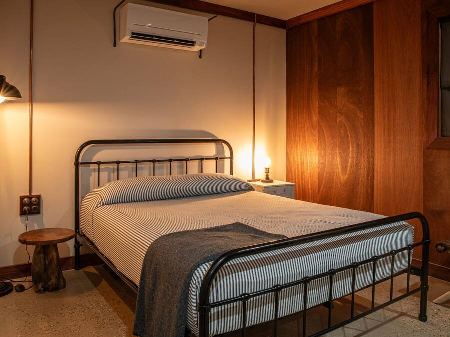 Una cama con estructura metálica, una lámpara de noche, aire acondicionado y un taburete pequeño de madera.