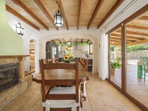 A dining area with a long wooden table, chairs, a stone fireplace, and a view of the kitchen and outdoor patio. - Son Bosc by Interhome (Inca)