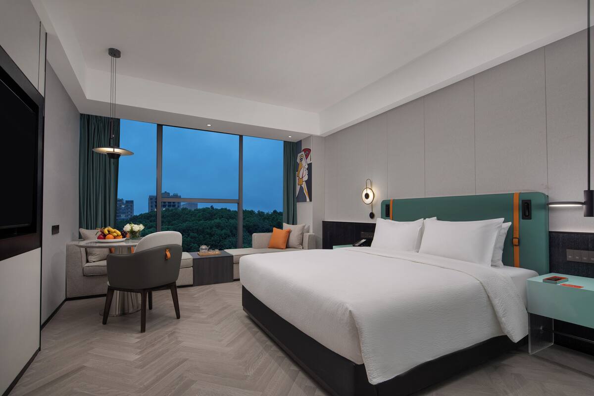 Ein modernes Hotelzimmer mit einem großen Bett, einem Schreibtisch mit Stuhl und Blick auf die Stadt durch große Fenster. Stadtansicht vom Fenster aus.