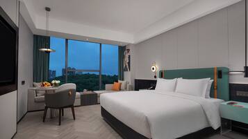 A modern hotel room with a large bed, a desk with a chair, a view of the cityscape through large windows, and a cityscape view from the window.