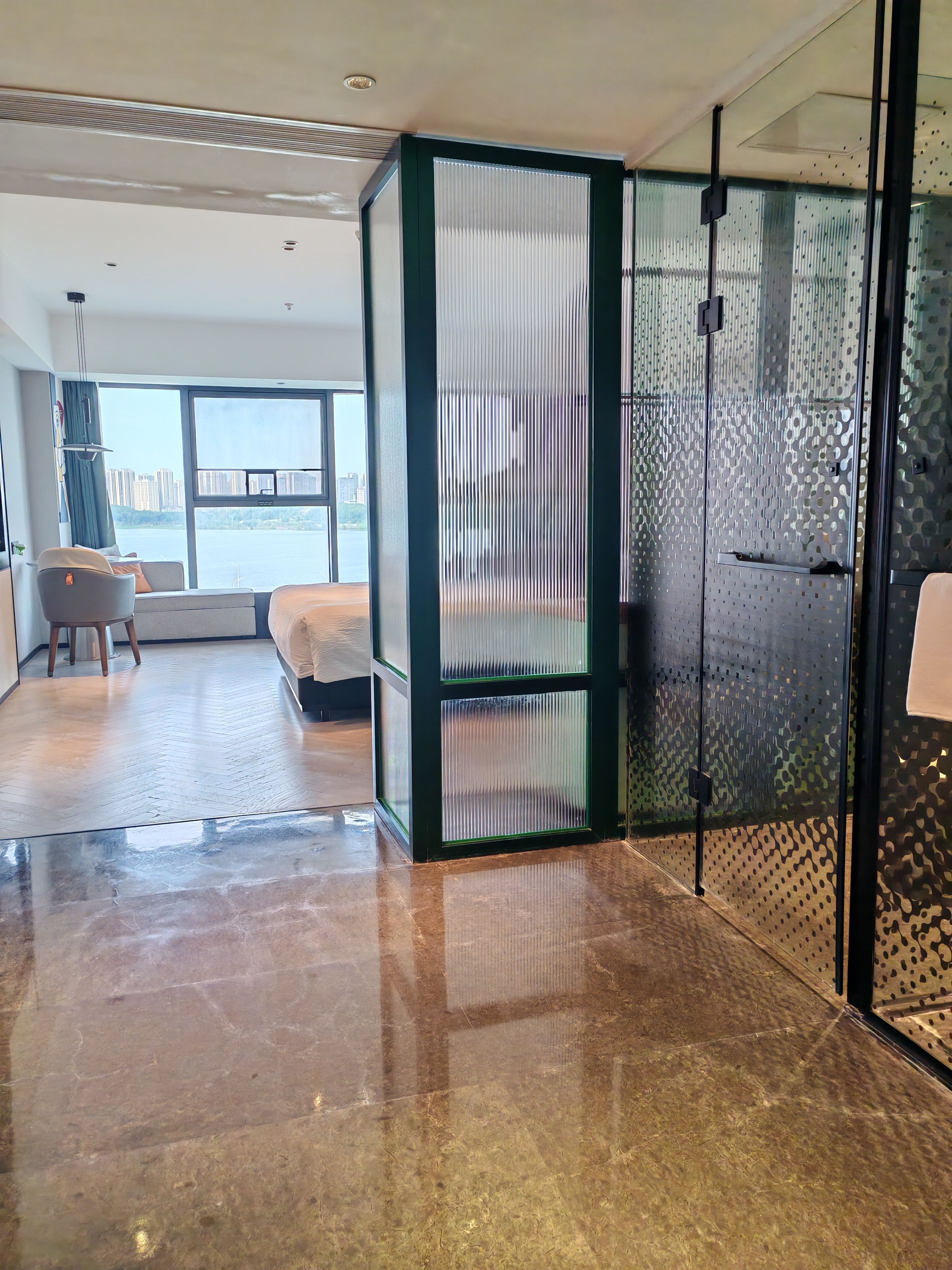 A hotel room with a large window, a bed, a seating area with a chair and a table, and a glass partition with a patterned design.