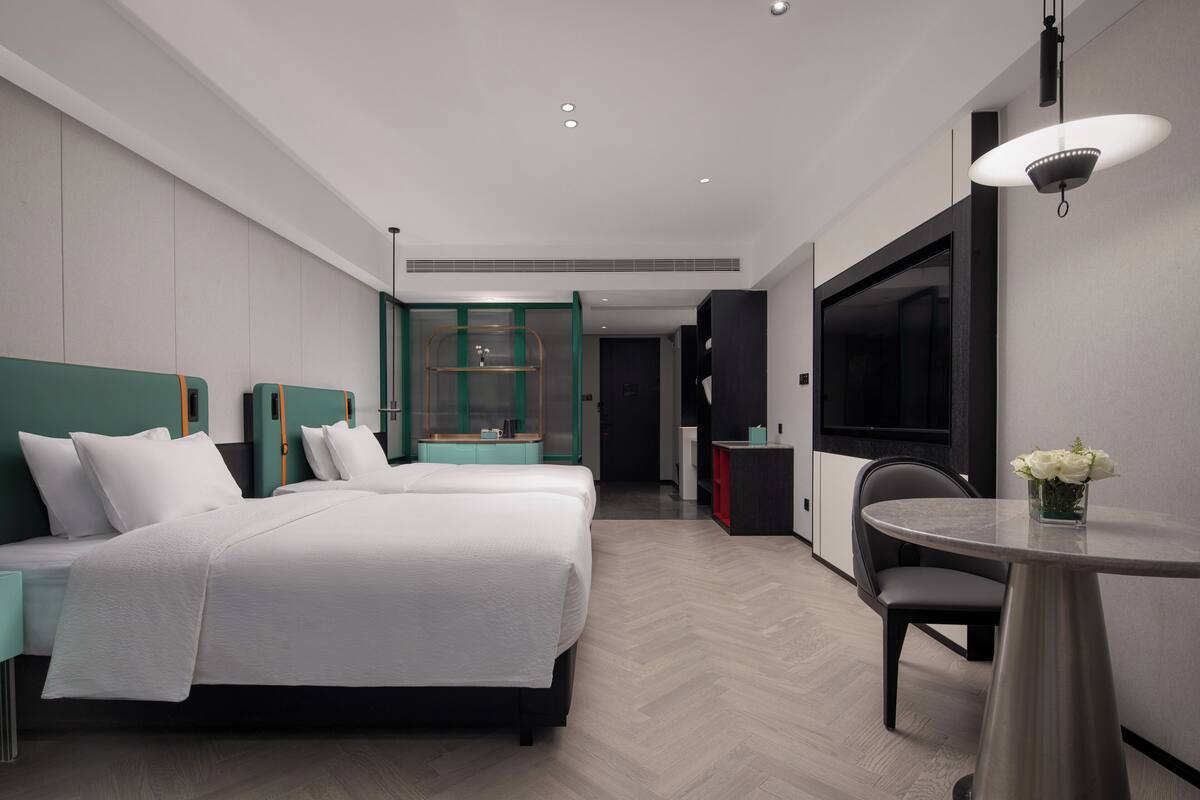 Ein modernes Hotelzimmer mit zwei Betten, einem Esstisch, einem Fernseher und einem sichtbaren Badezimmer im Hintergrund.