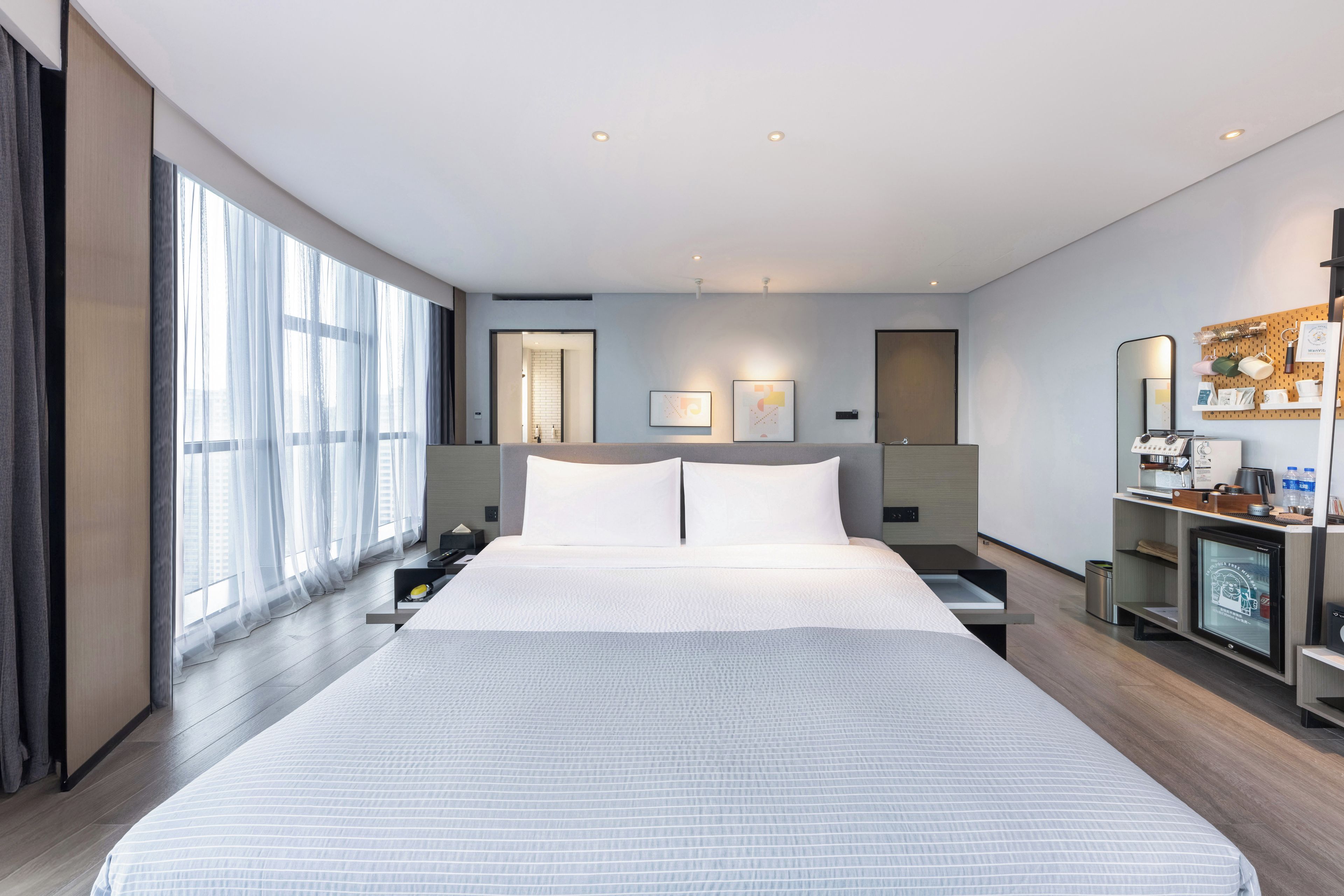 A modern hotel room with a large bed, a desk with a computer, and a bathroom visible through an open door.