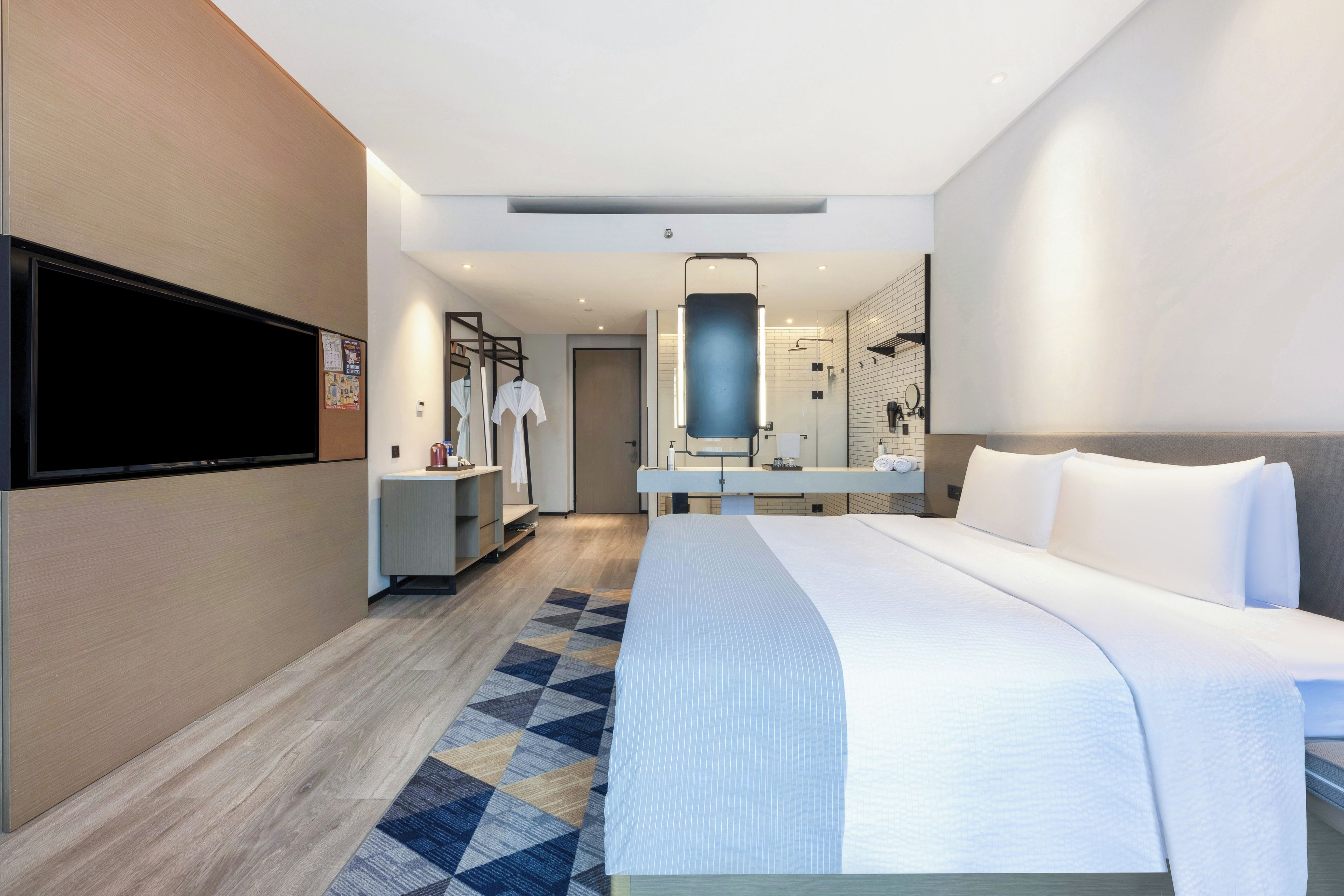 Una habitación de hotel moderna con una cama grande, un televisor de pantalla plana montado en la pared, un baño con espejo y lavamanos, y una pequeña zona de estar con una mesa y sillas.