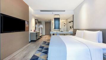 Una habitación de hotel moderna con una cama grande, un televisor de pantalla plana montado en la pared, un baño con espejo y lavamanos, y una pequeña zona de estar con una mesa y sillas.