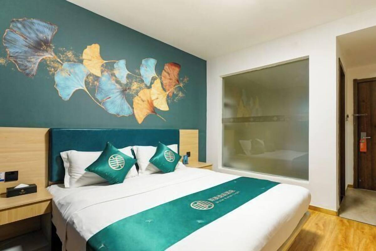 A hotel room with a bed, a nightstand, a large window, and a wall with a floral painting.