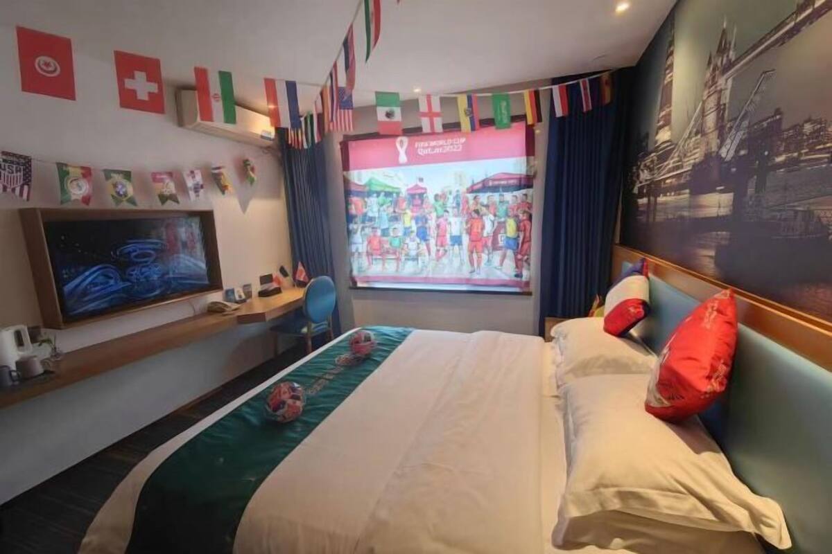 A hotel room with a bed, a TV, a desk, and a window with a view of a sports event.