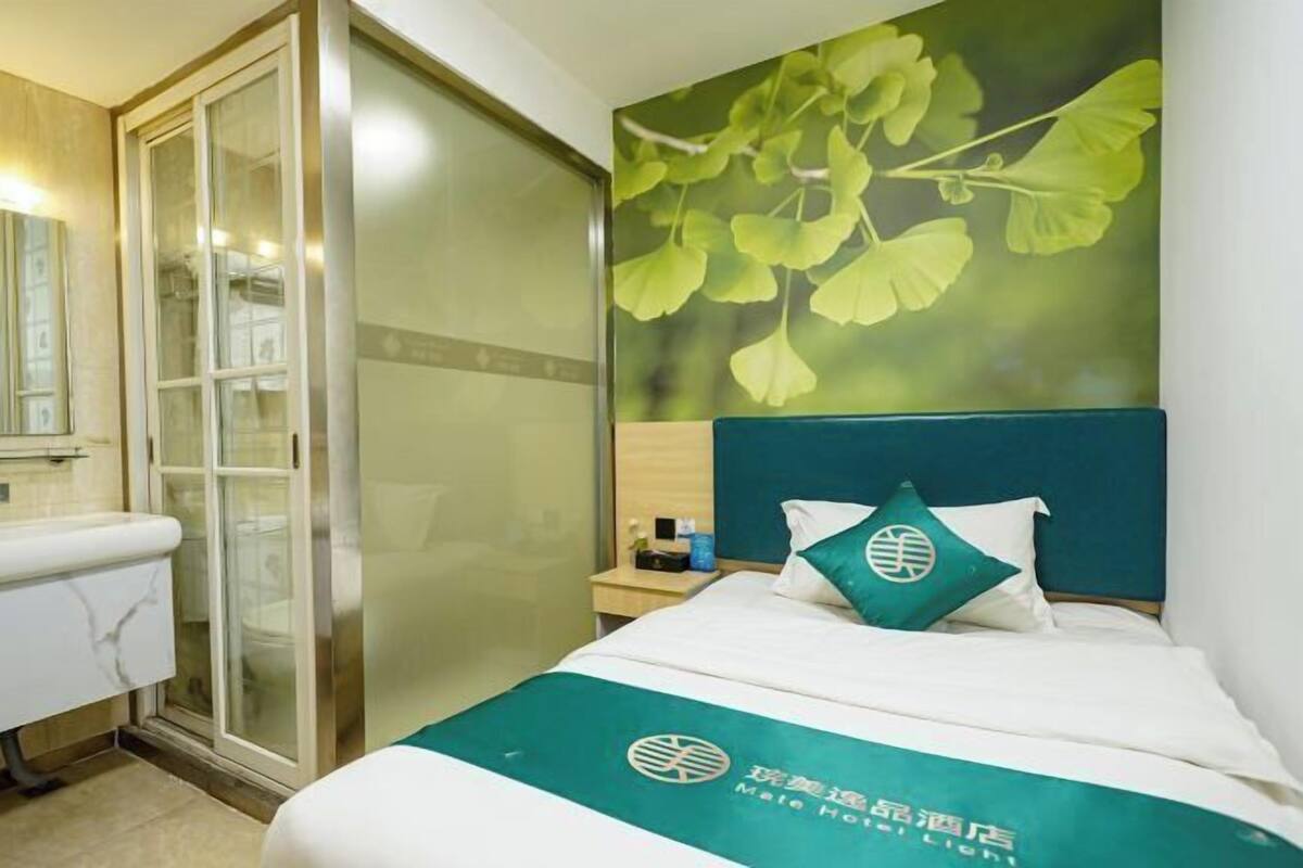 A hotel room with a bed, a nightstand, a bathroom with a toilet and a sink, and a large window with a view of trees.