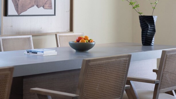 Un comedor moderno con una mesa larga, sillas de mimbre, un cuenco de fruta y un jarrón con plantas verdes.