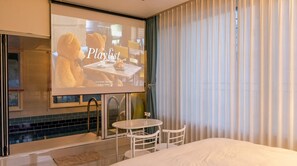 A hotel room with a bed, a small table, a chair, a TV screen displaying a movie, and a window with curtains.