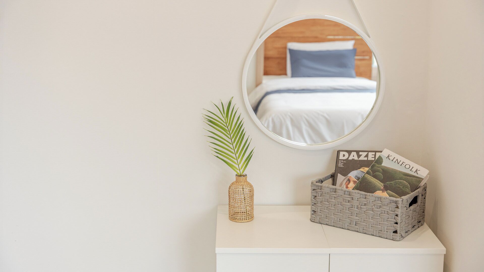 A bedroom with a bed, a mirror, a plant, and a wicker basket with magazines.