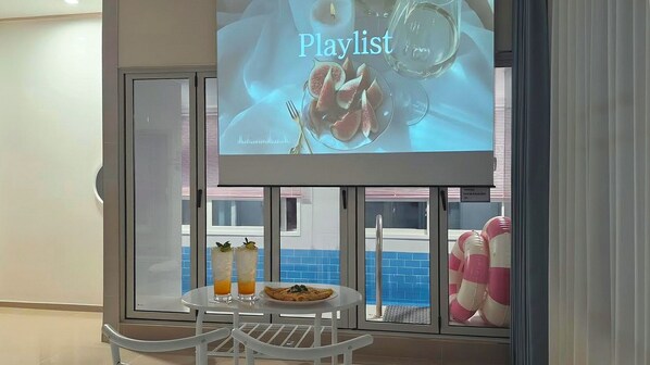 A room with a glass door, a table with a plate and drinks, and a projection screen displaying 'Playlist'.