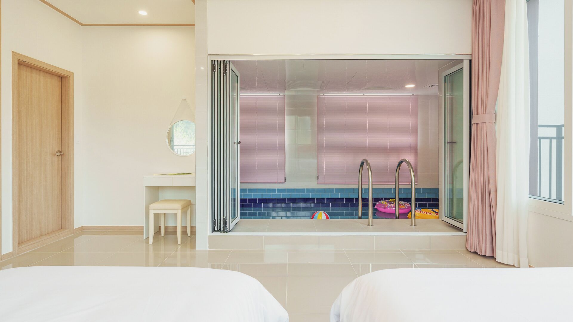 Une chambre d’hôtel avec deux lits, une salle de bain avec une porte en verre, un petit tabouret et une fenêtre avec des rideaux.