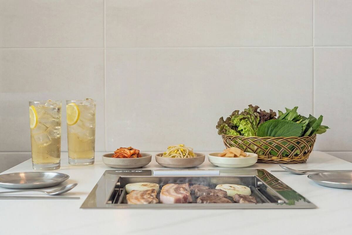Una mesa de comedor con dos vasos de té helado, fuentes de comida, una cesta de hojas verdes y una parrilla con carne.
