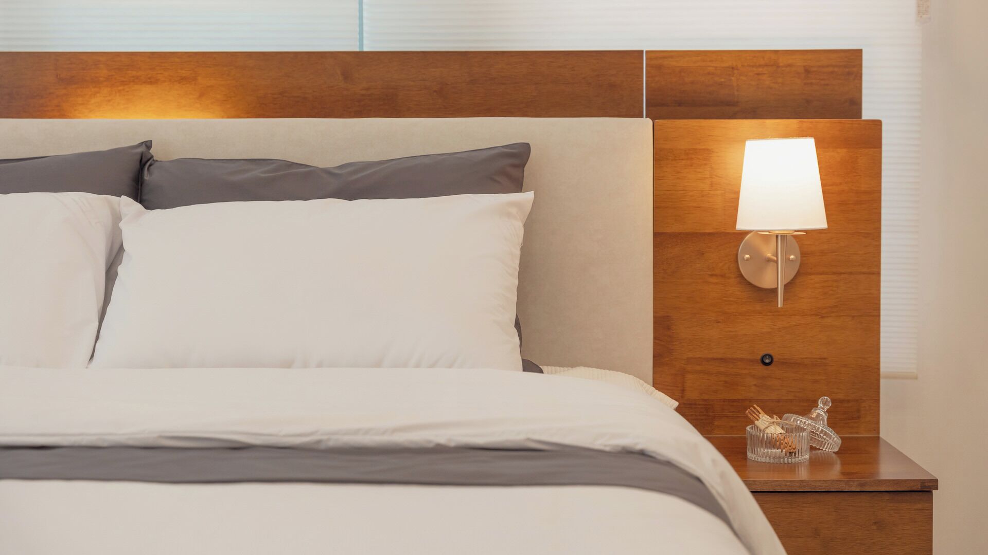 Una cama bien tendida con ropa de cama blanca y gris, cabecera de madera, lámpara de noche y un pequeño objeto decorativo en la mesita.