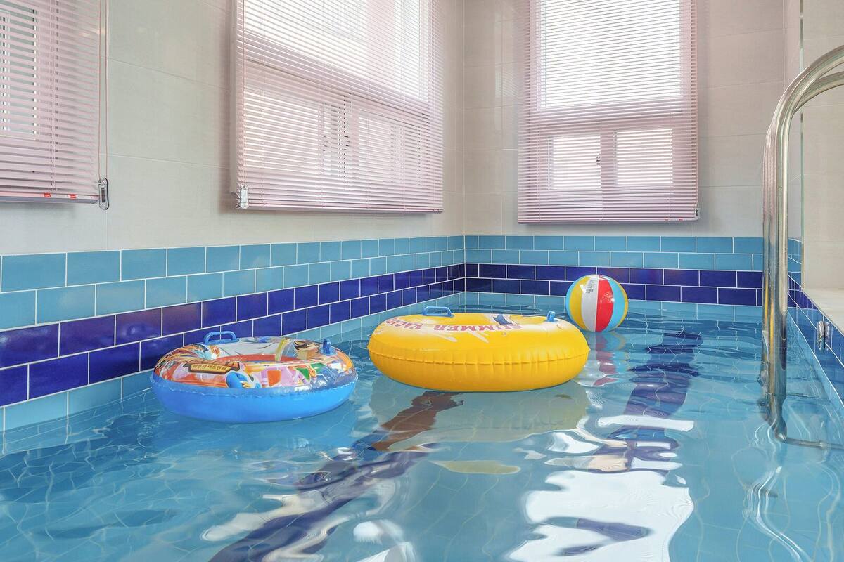 Una piscina interior con aros inflables azules y amarillos, una pelota de playa y paredes de azulejos.