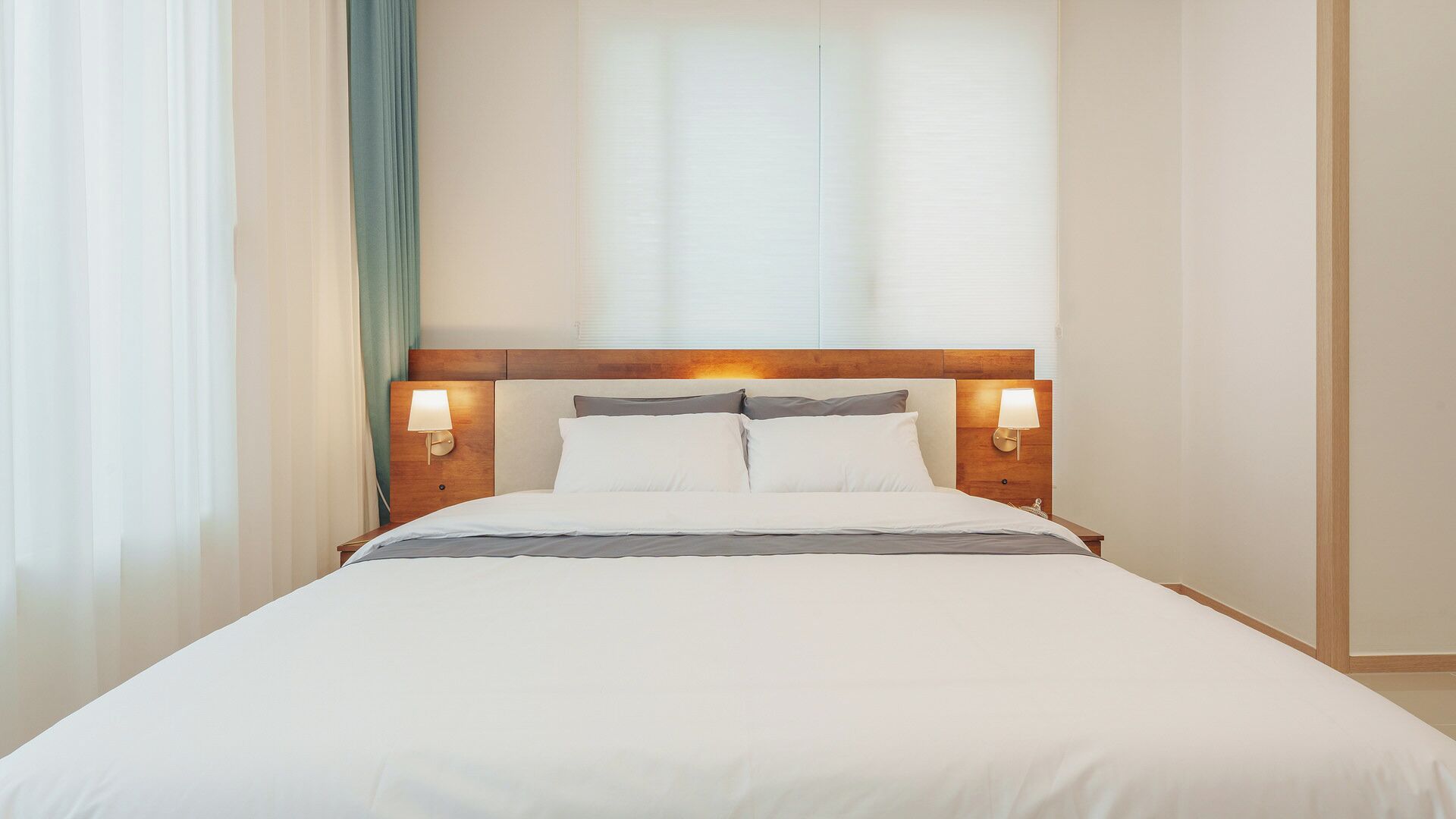 Une chambre d’hôtel avec un lit, deux lampes de chevet, une tête de lit en bois et des rideaux en voile.